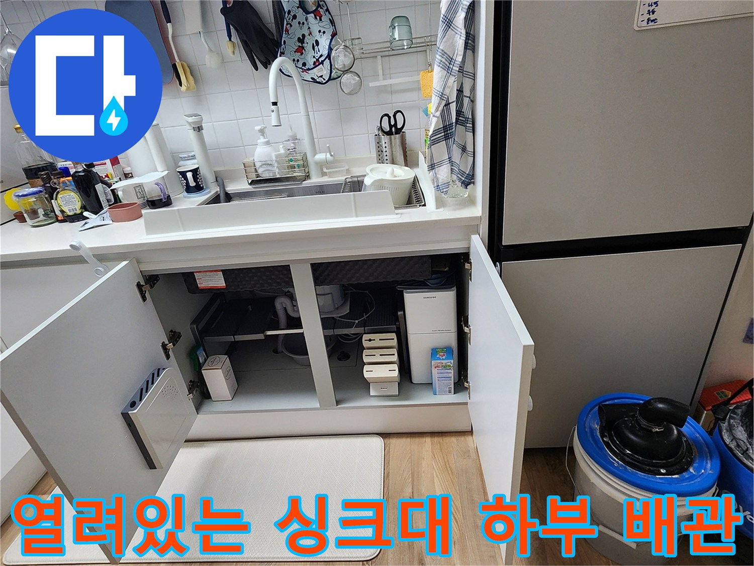 싱크대 아래 수납장이 열려 있는 배관 점검 현장 사진입니다.