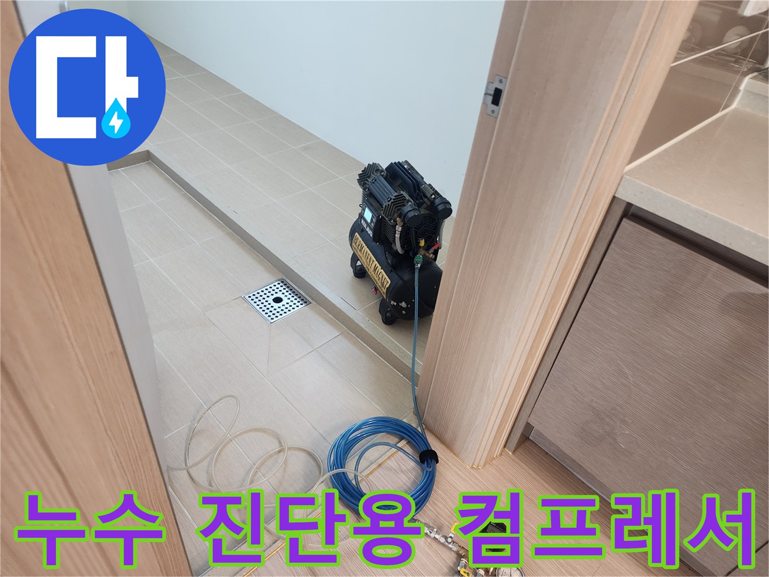 화장실 바닥 배수구 옆에 위치한 검정색 에어 컴프레서와 파란색 호스