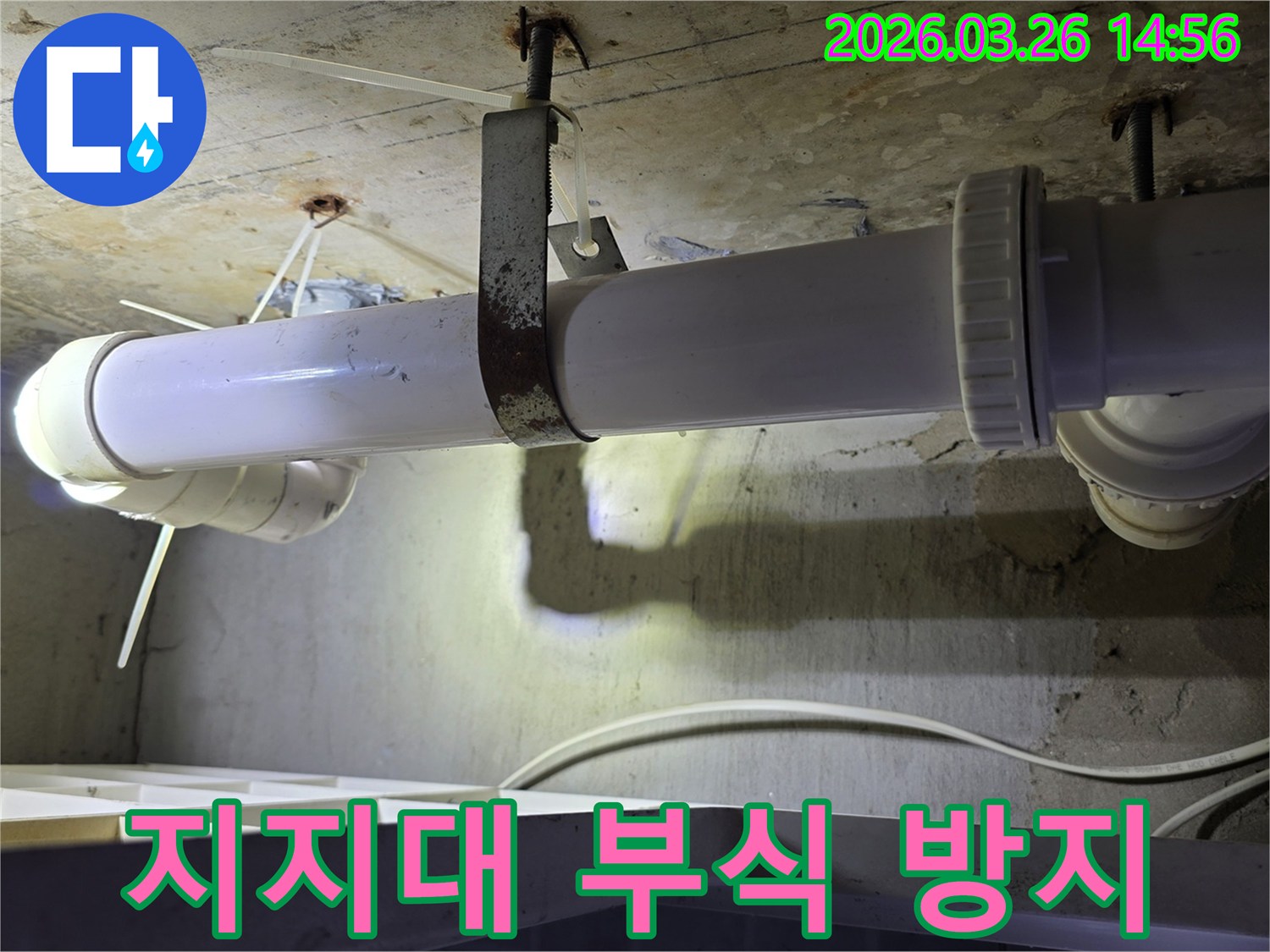 콘크리트 천장에 고정된-하얀색 배관과-심하게 녹슨 금속 지지대의 모습으로-부식으로 인한 누수 위험을 보여줍니다