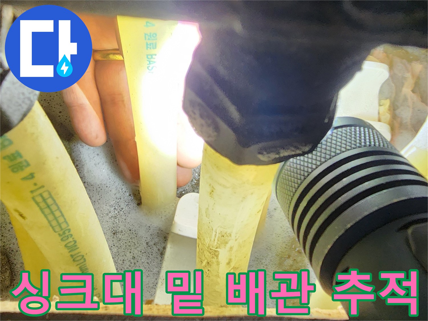 손전등을 사용하여-싱크대 하부장 내부의 흰색 배관들을 점검하는 모습