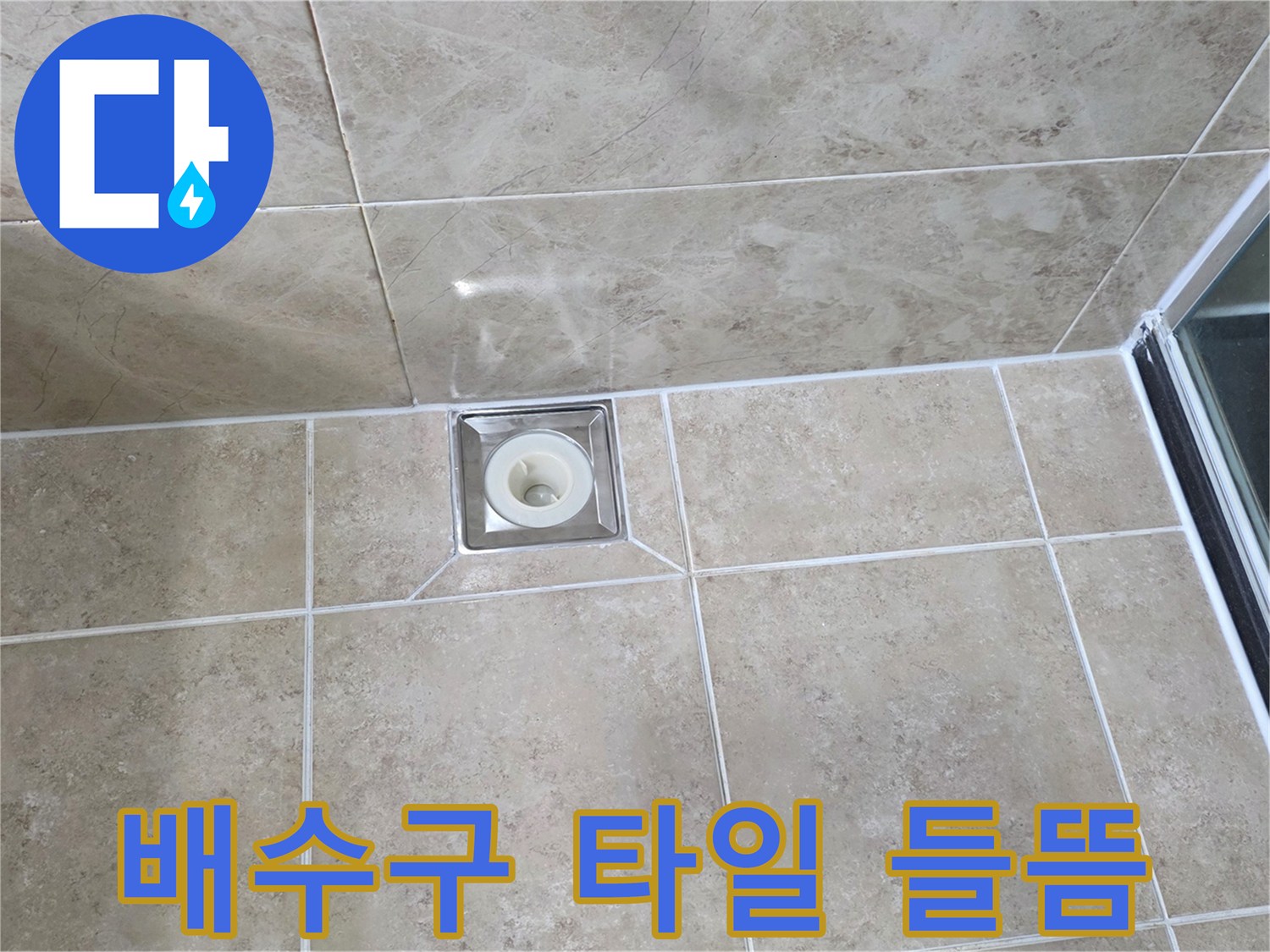 베이지색 타일 바닥에 위치한-사각형 금속 배수구의 모습으로-주변 타일의 접합부와-줄눈의 상태를 상세히 보여줍니다