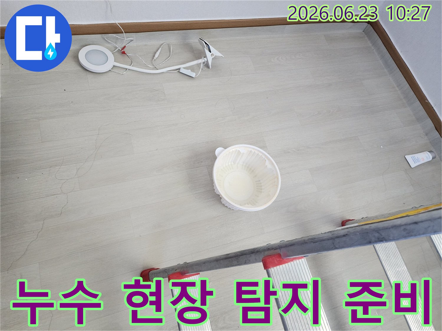 누수로 인해 바닥에 떨어진 물을 임시로 받는 그릇과-누수 지점 확인 및 수리를 위한 사다리-작업용 조명 장비가 바닥에 놓여 있는-누수 공사 전 현장 모습입니다