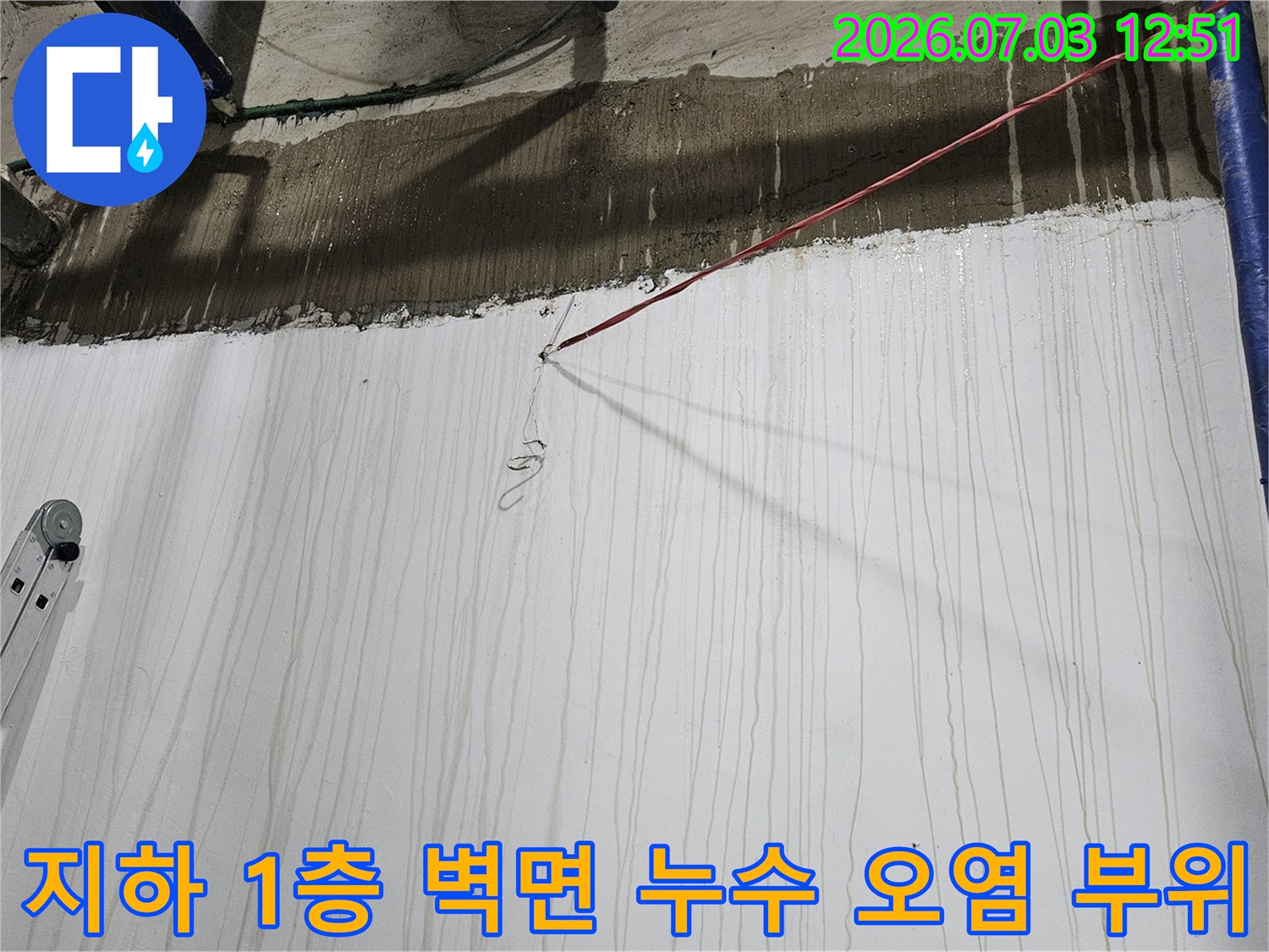 흰색으로 마감된 지하 벽면에 수직으로 형성된 갈색 누수 얼룩-상단 콘크리트 구조물과 파란 배관들