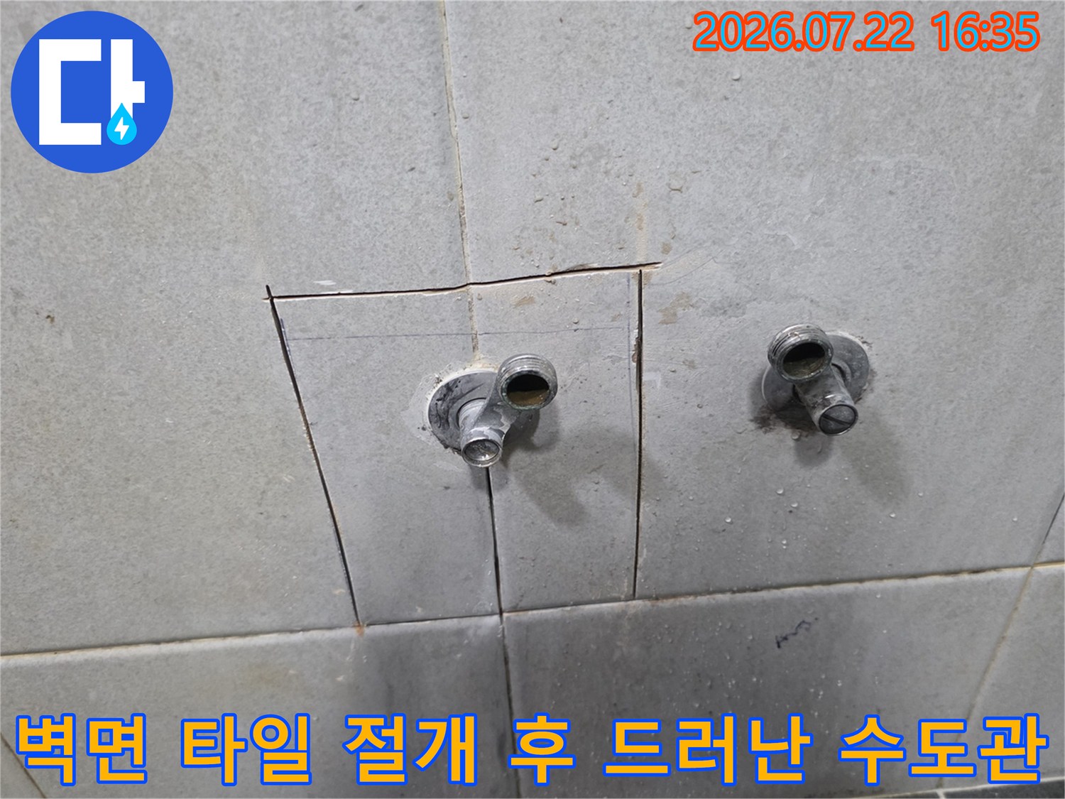 흰색 계열의 타일 벽면에 구멍이 뚫려 있고 그 사이로 금속제 배관 두 개가 돌출된 사진