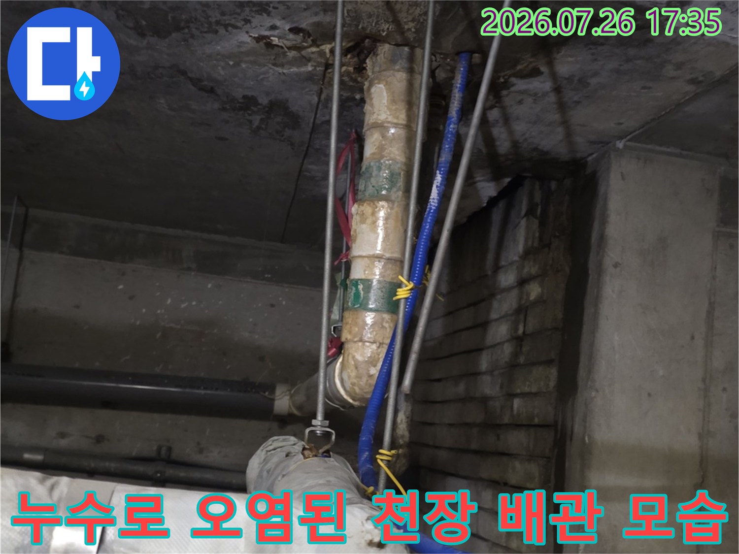 천장에 고정된 금속 지지대와 물때가 낀 흰색 배관 및 파란색 유연한 호스