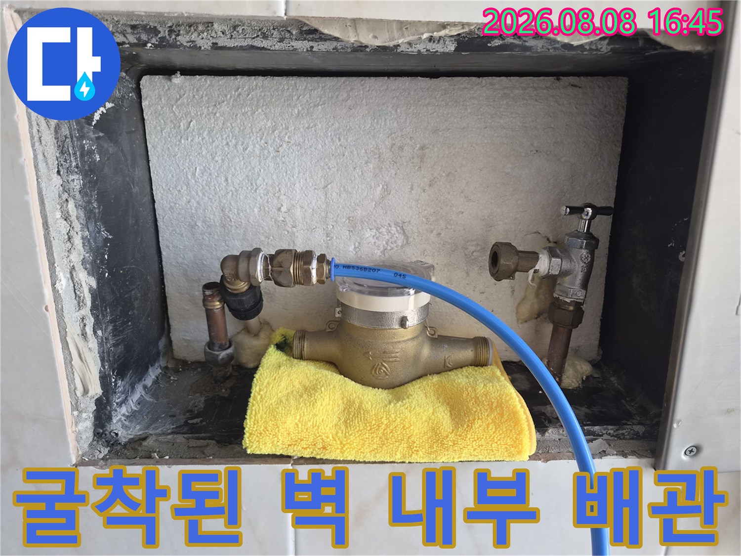 벽면 타일을 뜯어낸 공간에 드러난 금속 부속과 파란색 점검용 호스 모습입니다.