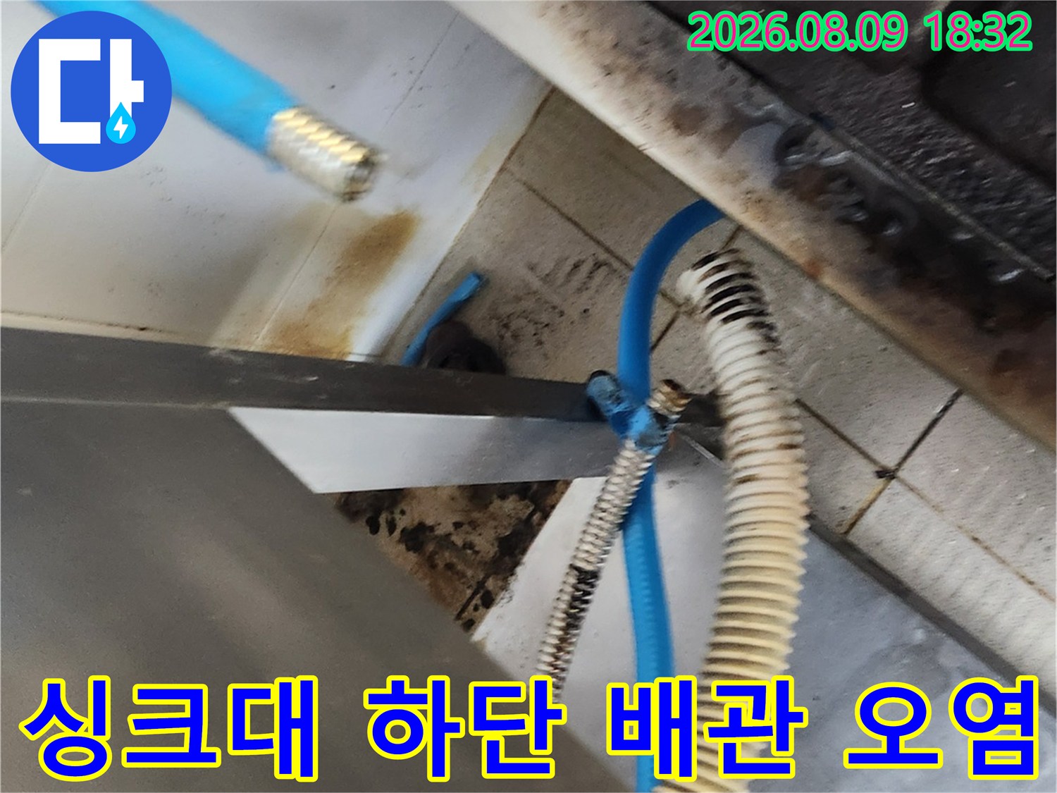 오염된 수도 부속과 파란색 호스가 보이는 싱크대 하부 모습입니다.