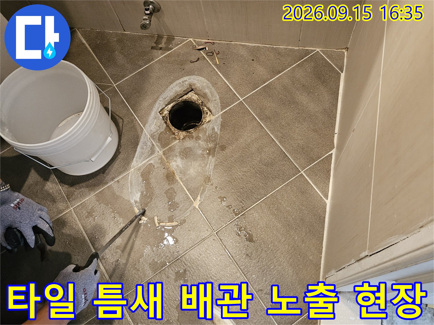 깨진 타일 사이로 드러난 하수구 배관과 주변 잔해물 사진입니다.